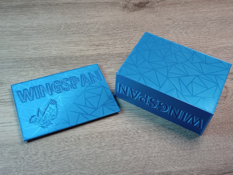 May include: Two-piece set of turquoise blue game storage boxes. The lid and the base of the larger box and the lid of the smaller box feature the word "WINGSPAN" and a bird graphic. Both boxes have a geometric pattern.