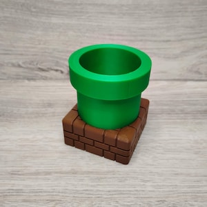 May include: A green pipe, reminiscent of a video game, sits atop a brown brick-like base. The pipe is cylindrical with a wide opening. The base is rectangular with a brick pattern. The objects are made of a solid material.
