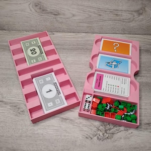 May include: Pink Monopoly game pieces and cards in a pink tray. The tray has slots for money, chance cards, and community chest cards. There are also slots for houses and hotels. The tray is open and the pieces are visible.