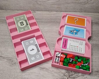 Puede incluir: Piezas y cartas de juego de Monopoly rosas en una bandeja rosa. La bandeja tiene ranuras para dinero, cartas de oportunidad y cartas de caja de la comunidad. También hay ranuras para casas y hoteles. La bandeja está abierta y las piezas son visibles.