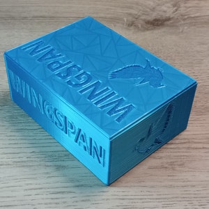 May include: A turquoise box with the word "WINGSPAN" embossed on the side. The box has a geometric pattern on the top and a bird silhouette. The box is made of a 3D printed material.