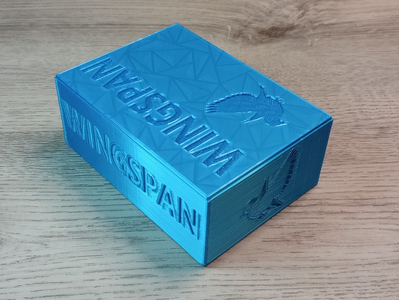 May include: A turquoise box with the word "WINGSPAN" embossed on the side. The box has a geometric pattern on the top and a bird silhouette. The box is made of a 3D printed material.