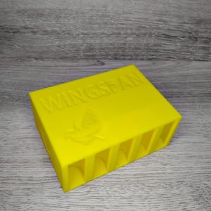 May include: Bright yellow rectangular box with the word "WINGSPAN" embossed on top, along with a bird emblem. The box has several vertical slots on one side. The item is likely a game organizer or storage container.