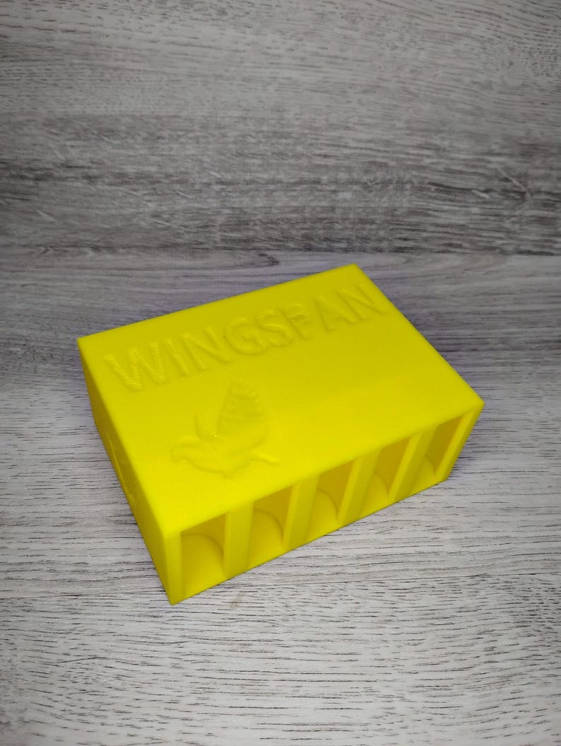 May include: Bright yellow rectangular box with the word "WINGSPAN" embossed on top, along with a bird emblem. The box has several vertical slots on one side. The item is likely a game organizer or storage container.