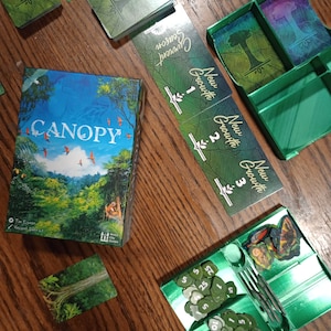 Könnte beinhalten: Ein grünes und blaues Brettspiel namens Canopy mit einer Schachtel, Karten und Spielsteinen. Die Schachtel hat ein grünes und blaues Design mit einem Bild einer Walddachkrone. Die Karten sind grün und haben Text, der "Neues Wachstum" und "Saison" sagt. Die Spielsteine sind grün und haben Zahlen darauf.