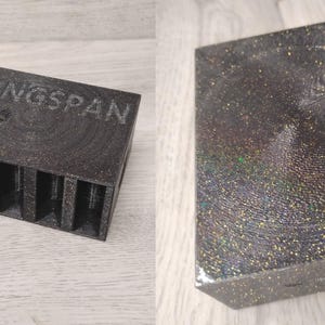 May include: A dark gray box with the word "WINGSPAN" and a bird graphic. The box has multiple vertical slots. The other item is a rectangular box with a glittery, iridescent surface.