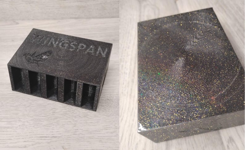 May include: A dark gray box with the word "WINGSPAN" and a bird graphic. The box has multiple vertical slots. The other item is a rectangular box with a glittery, iridescent surface.
