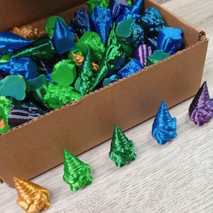 Op de afbeelding: Een doos gevuld met kleurrijke 3D-geprinte miniatuurboompjes. De boompjes zijn in verschillende tinten blauw, groen en goud. Er zijn vijf boompjes buiten de doos, in een rij gerangschikt.