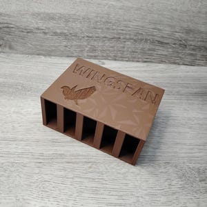 May include: A brown rectangular organizer with six vertical slots. The top surface features the word "WINGSPAN" and a bird graphic. The organizer is made of a solid material and is designed to hold small items.