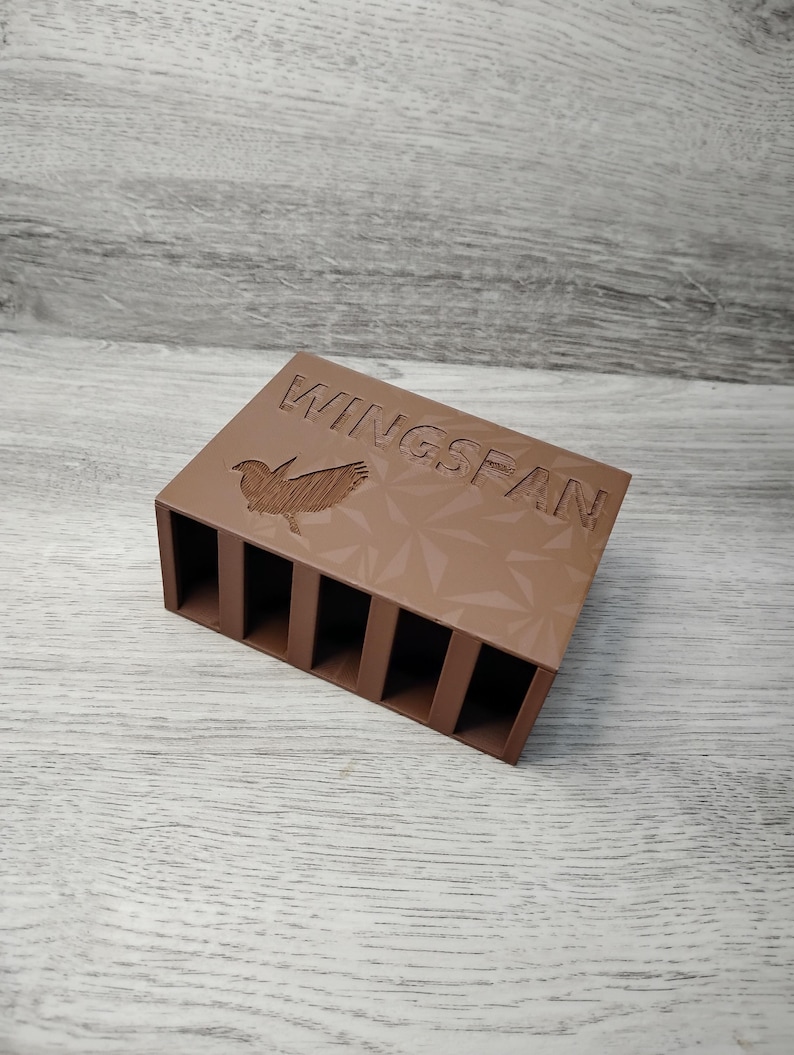 May include: A brown rectangular organizer with six vertical slots. The top surface features the word "WINGSPAN" and a bird graphic. The organizer is made of a solid material and is designed to hold small items.