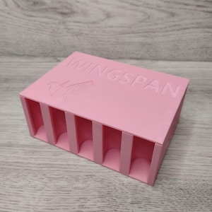 May include: A pink rectangular organizer with vertical dividers. The word "WINGSPAN" is embossed on the top, along with a bird graphic. The organizer is designed to hold small items.