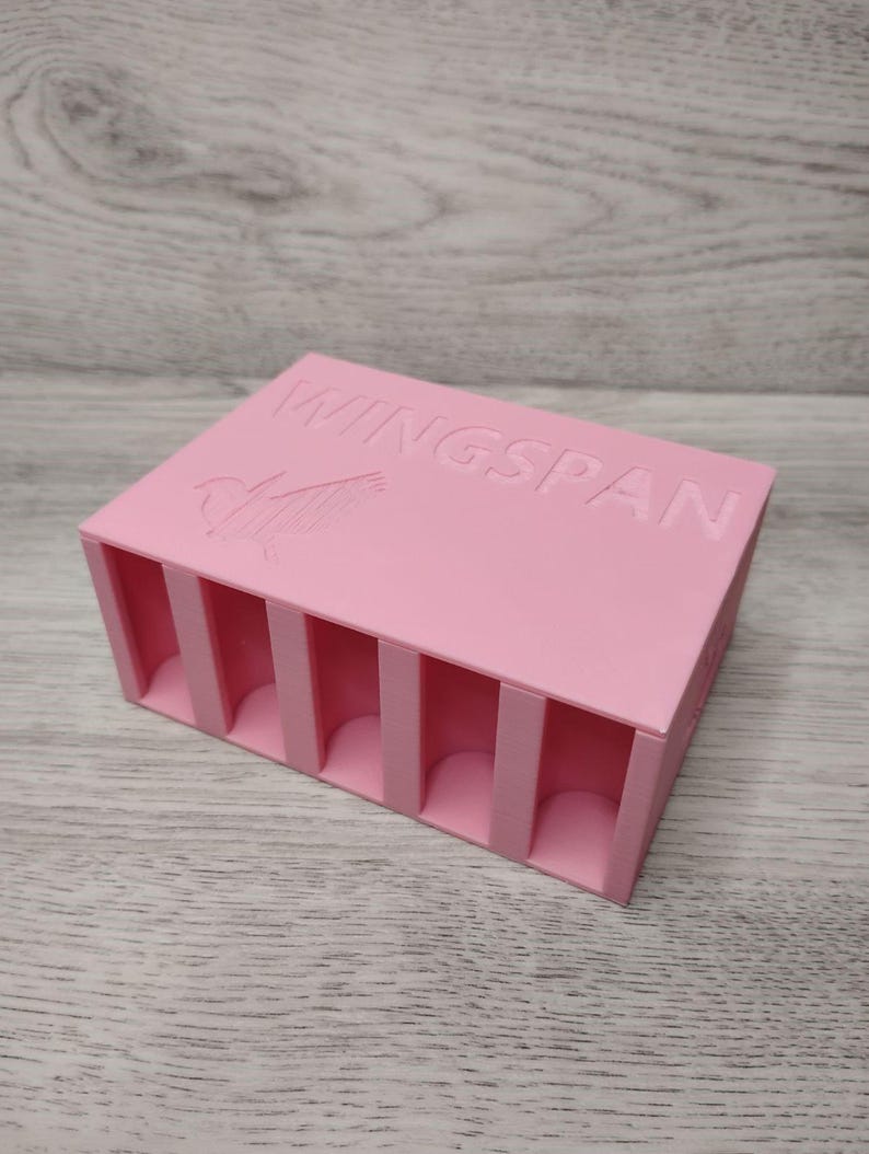 May include: A pink rectangular organizer with vertical dividers. The word "WINGSPAN" is embossed on the top, along with a bird graphic. The organizer is designed to hold small items.
