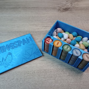May include: A blue Wingspan board game organizer. The organizer has compartments for game tokens and eggs. The lid is also blue and features the game's name and a bird illustration. The tokens are colorful, and the eggs are various pastel colors.