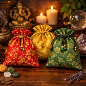 以下が含まれることがあります： 赤、金、緑の3つの装飾用巾着袋。それぞれに花柄とビーズのアクセントが付いています。袋は、背景にろうそくやその他の精神的なアイテムがある木製の表面に配置されています。