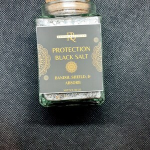May include: A glass jar with a cork lid filled with black salt. The label reads "Protection Black Salt" and "Banish, Shield, & Absorb". The jar is 10 ounces.
