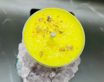 May include: A bright yellow tea light candle with glitter and dried herbs on top, set on a purple and white crystal. The candle is in a clear plastic container. The background is a dark, reflective surface.