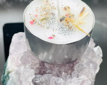 May include: A small, white candle in a silver metal container, adorned with dried herbs, glitter, and small pink accents. The candle sits atop a cluster of purple amethyst crystals, creating a natural and decorative aesthetic.