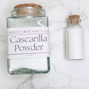 May include: Two glass jars with cork lids filled with white powder. The larger jar has a label that reads "Roots Botanica Co Cascarilla Powder 3.4 oz".