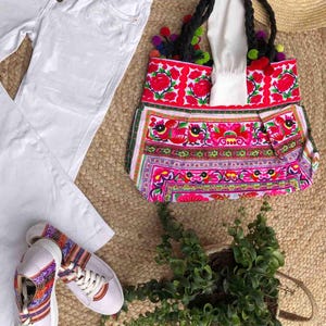K&ouml;nnte beinhalten: Eine farbenfrohe Tragetasche mit floralem Design, wei&szlig;e Jeans und wei&szlig;e Sneaker mit gemusterten Akzenten werden auf einer gewebten Matte pr&auml;sentiert. Die Tragetasche hat schwarze geflochtene Griffe und Pompons.