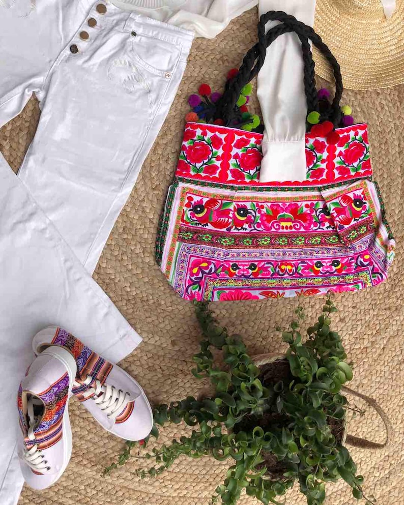 K&ouml;nnte beinhalten: Eine farbenfrohe Tragetasche mit floralem Design, wei&szlig;e Jeans und wei&szlig;e Sneaker mit gemusterten Akzenten werden auf einer gewebten Matte pr&auml;sentiert. Die Tragetasche hat schwarze geflochtene Griffe und Pompons.