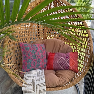 May include: A hanging wicker egg chair with a tan cushion, featuring a square pillow with a pink and teal geometric pattern, a rectangular pillow with a red and purple diamond design, and a white throw blanket with a brown geometric pattern.