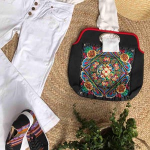K&ouml;nnte beinhalten: Draufsicht auf ein Mode-Ensemble mit wei&szlig;er Jeans, wei&szlig;er Bluse, schwarzen Sneakers mit farbenfrohen Mustern und einer schwarzen Handtasche mit lebendiger Stickerei. Ein Strohhut und eine kleine Topfpflanze vervollst&auml;ndigen den Look.