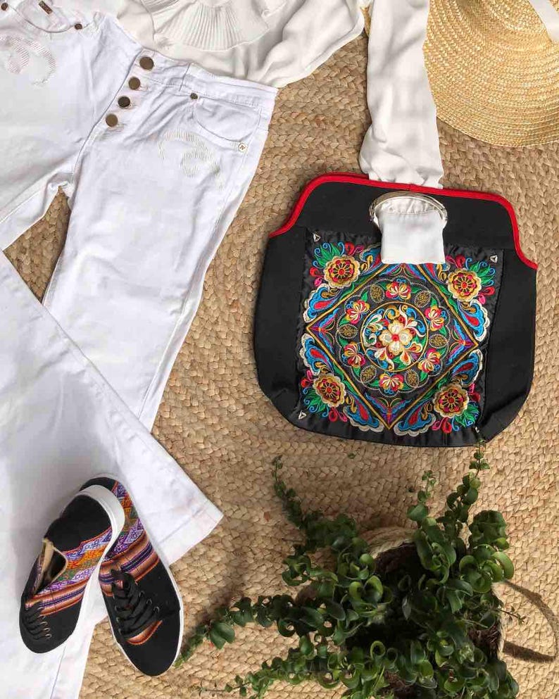 K&ouml;nnte beinhalten: Draufsicht auf ein Mode-Ensemble mit wei&szlig;er Jeans, wei&szlig;er Bluse, schwarzen Sneakers mit farbenfrohen Mustern und einer schwarzen Handtasche mit lebendiger Stickerei. Ein Strohhut und eine kleine Topfpflanze vervollst&auml;ndigen den Look.