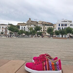 K&ouml;nnte beinhalten: Ein Paar leuchtend pinkfarbene Sneaker mit wei&szlig;en Sohlen und roten Schn&uuml;rsenkeln. Die Seiten der Schuhe weisen ein farbenfrohes Webmuster mit geometrischen Designs auf. Die Schuhe stehen vor einem Stadtplatz mit Geb&auml;uden und einem bew&ouml;lkten Himmel.