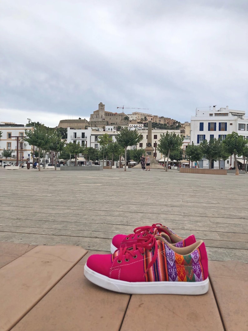 K&ouml;nnte beinhalten: Ein Paar leuchtend pinkfarbene Sneaker mit wei&szlig;en Sohlen und roten Schn&uuml;rsenkeln. Die Seiten der Schuhe weisen ein farbenfrohes Webmuster mit geometrischen Designs auf. Die Schuhe stehen vor einem Stadtplatz mit Geb&auml;uden und einem bew&ouml;lkten Himmel.