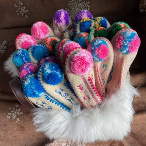 May include: A group of white felt slippers with colorful pom-poms and embroidered designs. The slippers are arranged in a basket lined with white fur.