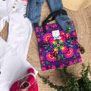 K&ouml;nnte beinhalten: Eine farbenfrohe Tragetasche mit einem floralen Design in Pink, Blau und Gr&uuml;n, mit einer Jeansjacke, einer wei&szlig;en Hose und pinkfarbenen Turnschuhen. Die Tasche hat einen schwarzen Riemen und Holzperlen. Ein Strohhut ist ebenfalls sichtbar.
