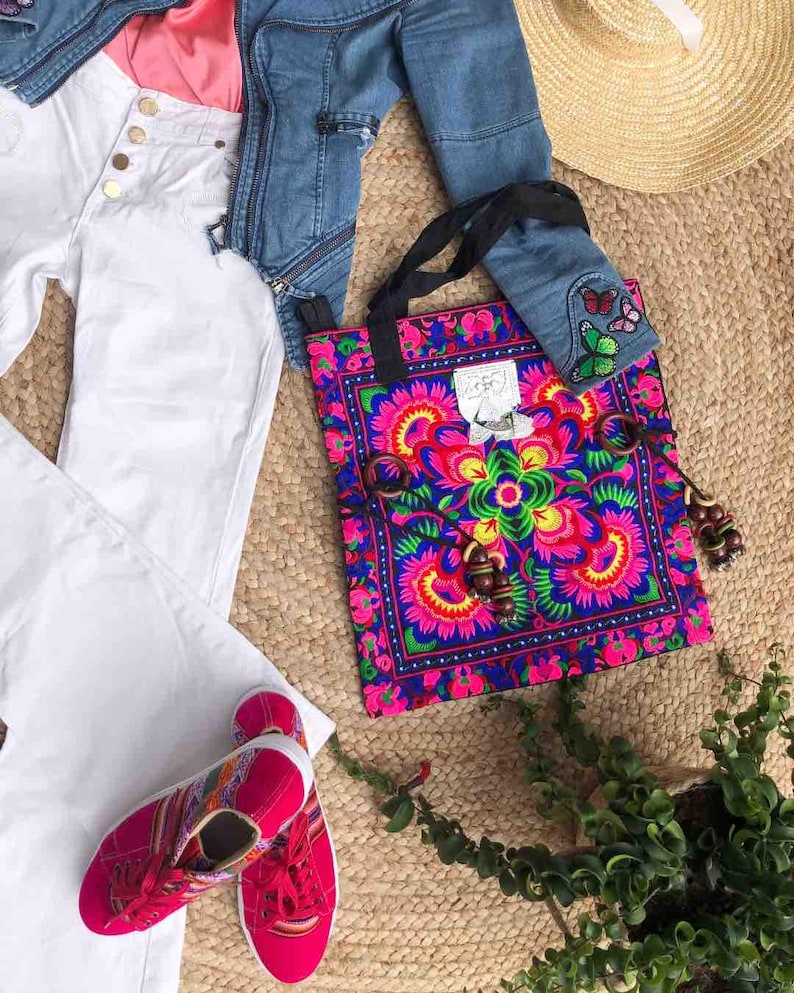 K&ouml;nnte beinhalten: Eine farbenfrohe Tragetasche mit einem floralen Design in Pink, Blau und Gr&uuml;n, mit einer Jeansjacke, einer wei&szlig;en Hose und pinkfarbenen Turnschuhen. Die Tasche hat einen schwarzen Riemen und Holzperlen. Ein Strohhut ist ebenfalls sichtbar.