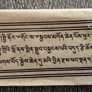 May include: A rectangular, light brown paper with black text and a black border. The text is in a foreign language. The paper is folded at the top and bottom. The paper appears to be aged.
