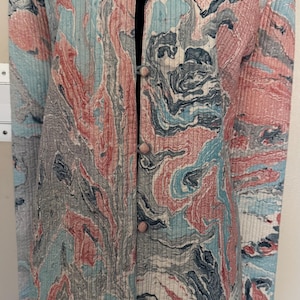 May include: A long-sleeved, ribbed jacket featuring a marbled pattern in shades of light blue, dusty pink, and charcoal gray. The garment has a mandarin collar and a front button closure with four small, round, matching fabric-covered buttons.
