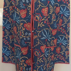 May include: A long-sleeved quilted jacket in a deep blue hue, featuring a vibrant floral and leaf pattern in shades of red, light blue, and yellow. The garment includes a mandarin collar, pink piping trim, and a front closure with fabric-covered buttons.