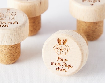 May include: Four wooden bottle stoppers with different designs. One stopper has a Christmas tree and the words "Joyeux Noel Papi". Another stopper has a wine glass, wine bottle, and the words "Feaux Noel Papi". A third stopper has the words "Joyeux Noel Papi". The fourth stopper has a cartoon pug with antlers and the words "Pour mon Papi chéri".