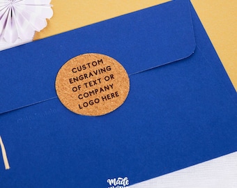 May include: A circular cork sticker featuring the black engraved text CUSTOM ENGRAVING OF TEXT OR COMPANY LOGO HERE, adhered to the flap of a vibrant blue envelope. The sticker has a textured, natural brown surface and a clean, circular edge.