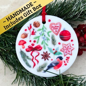May include: White ceramic Christmas ornament with a red ribbon and a variety of festive illustrations, including a red bird, holly, berries, a star, and a gingerbread heart that says "Frohes Fest".