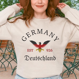Peut inclure: Sweat-shirt crème avec le mot "GERMANY" en arc de cercle et "Deutschland" en dessous. Un aigle stylisé aux ailes noires et rouges est centré. Le texte "EST 926" est également présent.