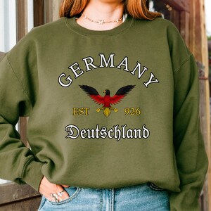 Peut inclure: Sweat-shirt vert olive avec le mot "GERMANY" en arc de cercle et "Deutschland" en dessous. Un emblème d'aigle rouge et noir est centré, avec "EST 1926" et "926" de chaque côté. Le sweat-shirt est porté avec un jean bleu.