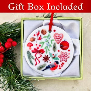 May include: A white ceramic Christmas ornament with a red ribbon and a variety of festive designs, including a red bird, a star, a heart, and a mushroom. The ornament is in a white gift box with a green lid. The text "Frohes Fest" is visible on the ornament.