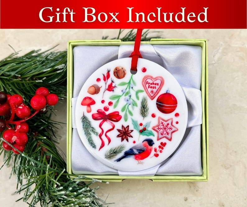 May include: A white ceramic Christmas ornament with a red ribbon and a variety of festive designs, including a red bird, a star, a heart, and a mushroom. The ornament is in a white gift box with a green lid. The text "Frohes Fest" is visible on the ornament.