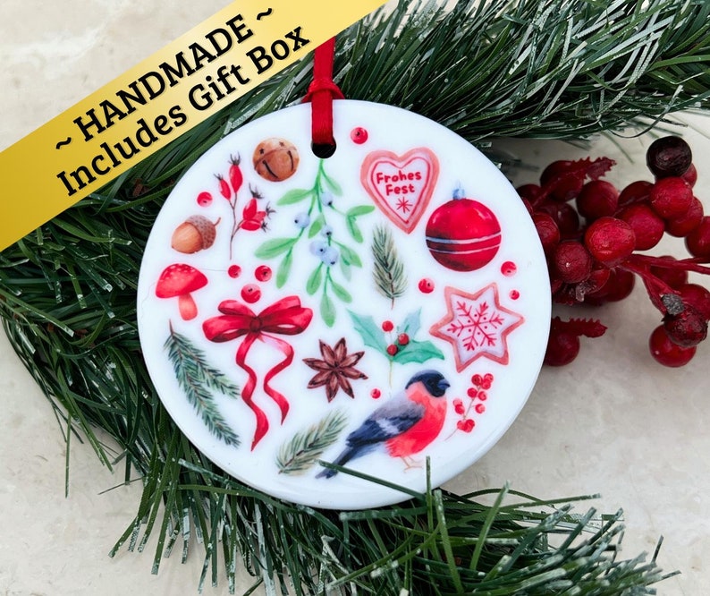 May include: White ceramic Christmas ornament with a red ribbon and a variety of festive illustrations, including a red bird, holly, berries, a star, and a gingerbread heart that says "Frohes Fest".