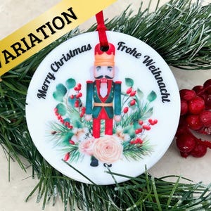 May include: White round Christmas ornament with a nutcracker illustration, green foliage, and pink roses. The ornament has the text "Merry Christmas" and "Frohe Weihnachten" on it.