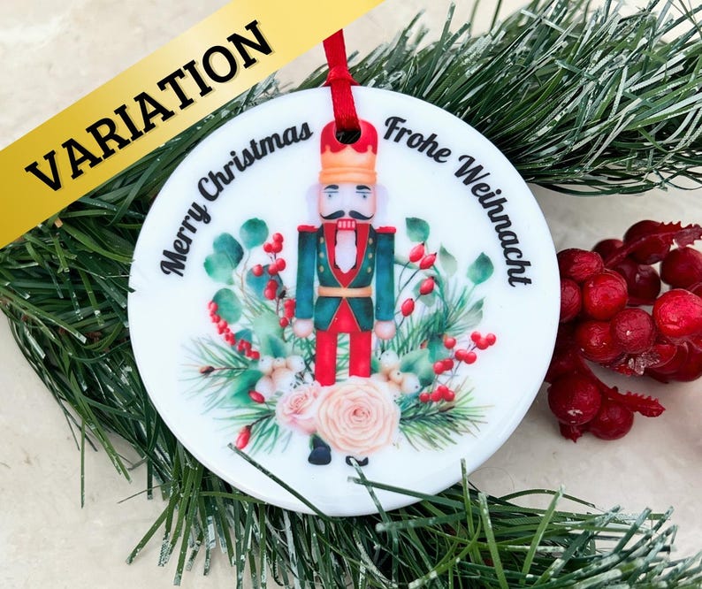 May include: White round Christmas ornament with a nutcracker illustration, green foliage, and pink roses. The ornament has the text "Merry Christmas" and "Frohe Weihnachten" on it.