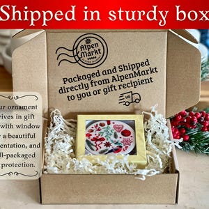May include: A brown cardboard box with a window showing a round Christmas ornament with a red, white, and green design. The box is filled with white shredded paper. The box has a label that says "Packaged and Shipped directly from AlpenMarkt to you or gift recipient".