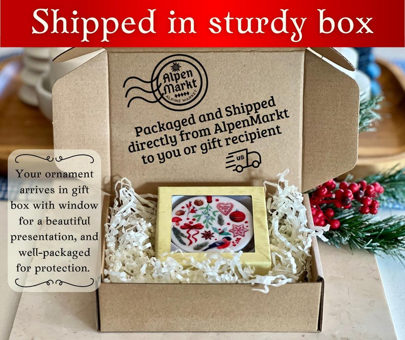 May include: A brown cardboard box with a window showing a round Christmas ornament with a red, white, and green design. The box is filled with white shredded paper. The box has a label that says "Packaged and Shipped directly from AlpenMarkt to you or gift recipient".