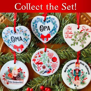 May include: Set of six white ceramic Christmas ornaments with various designs.  The ornaments are heart-shaped and round.  The designs include a wreath with the word "OPA", a wreath with the word "OMA", a design with the words "Ich Liebe Dich", a design with the words "Frohe Weihnachten", a design with the words "Merry Christmas", and a design with the words "Frohe Weihnachten" and a nutcracker.