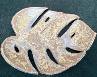 May include: A ceramic dish shaped like a monstera leaf. The dish is glazed in a light brown color with a speckled, iridescent finish.