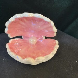 May include: A pink ceramic clam shell with a white pearl inside. The shell has a glossy finish and a slightly iridescent sheen.
